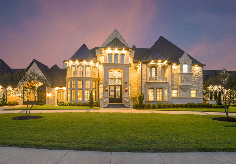 Luxurious stone mansion at dusk with warm exterior lighting and manicured lawn.